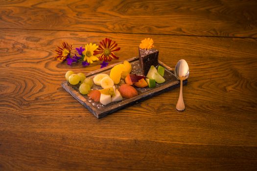 Assiette de fruits frais colorés avec un gâteau fondant au chocolat et poire, décorée de fleurs sur une table en bois.